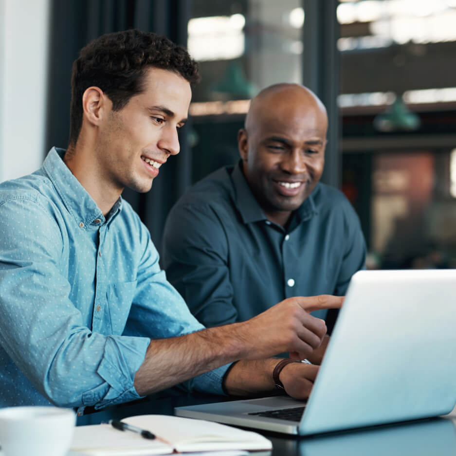 Two men collaborating in a breakout area in an office. One man is pointing to something on his laptop.