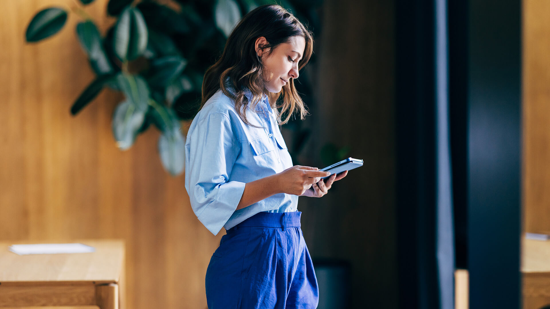 A woman stands indoors looking down at a tablet while holding it with both hands.