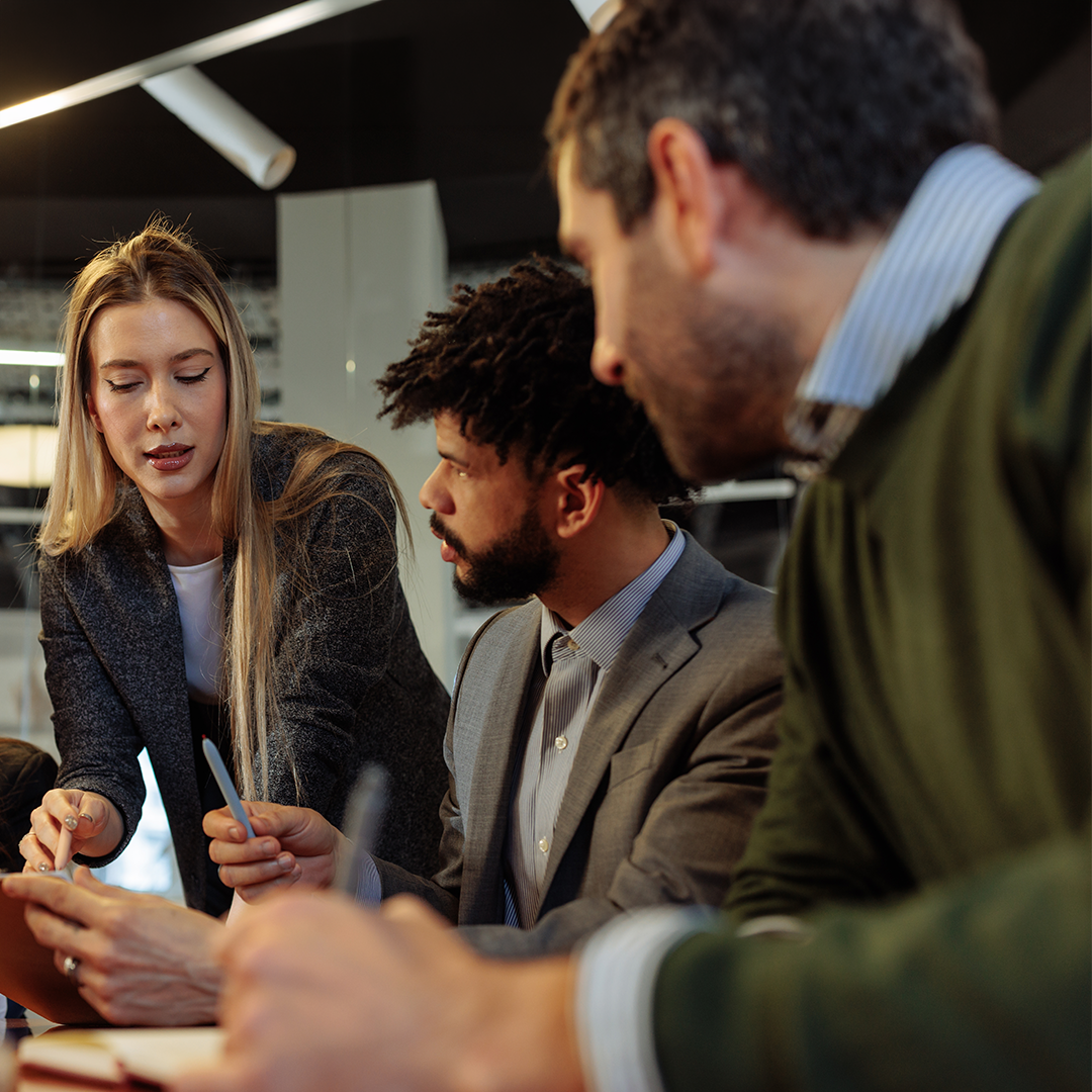 Three professionals are gathered around a desk in an office setting. A woman is standing and pointing at a document or tablet while two seated men look on. This provides a factual and neutral description without inferring unseen intent or emotion. It is designed to clearly explain the image to someone who cannot see it and avoids jargon.