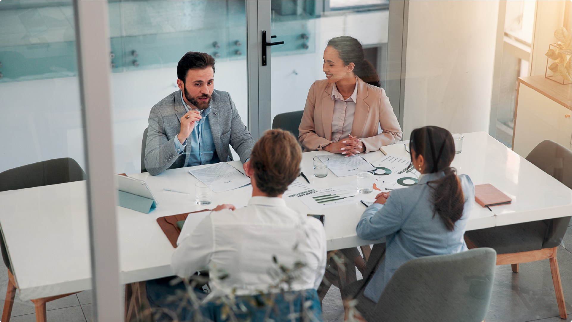 Four people are sitting around a white conference table in a glass-walled office. They are looking at documents and charts spread across the table while one man gestures with his hand.